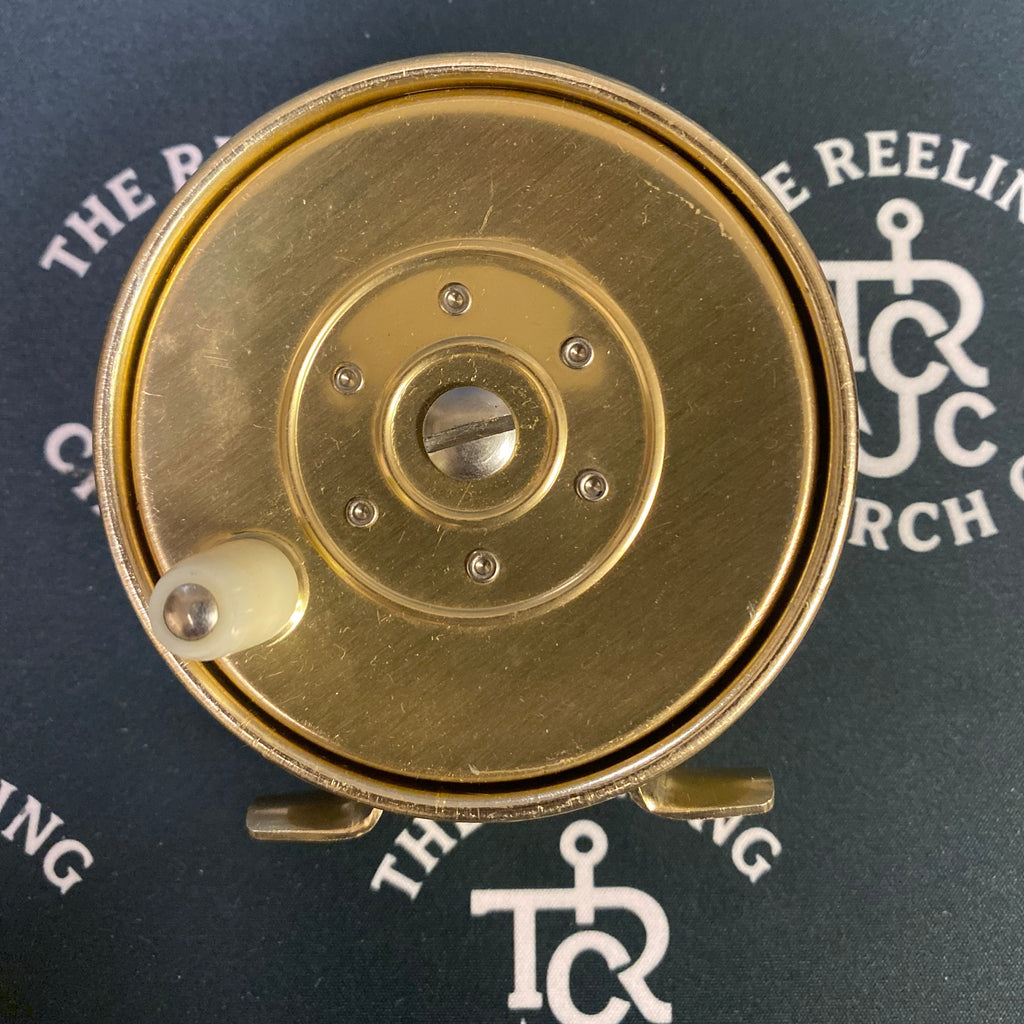 A close-up top-down view of a brass fishing reel with a small white plastic spool tip, set against a dark background with repeating white 'TRC Church Co.' logos.