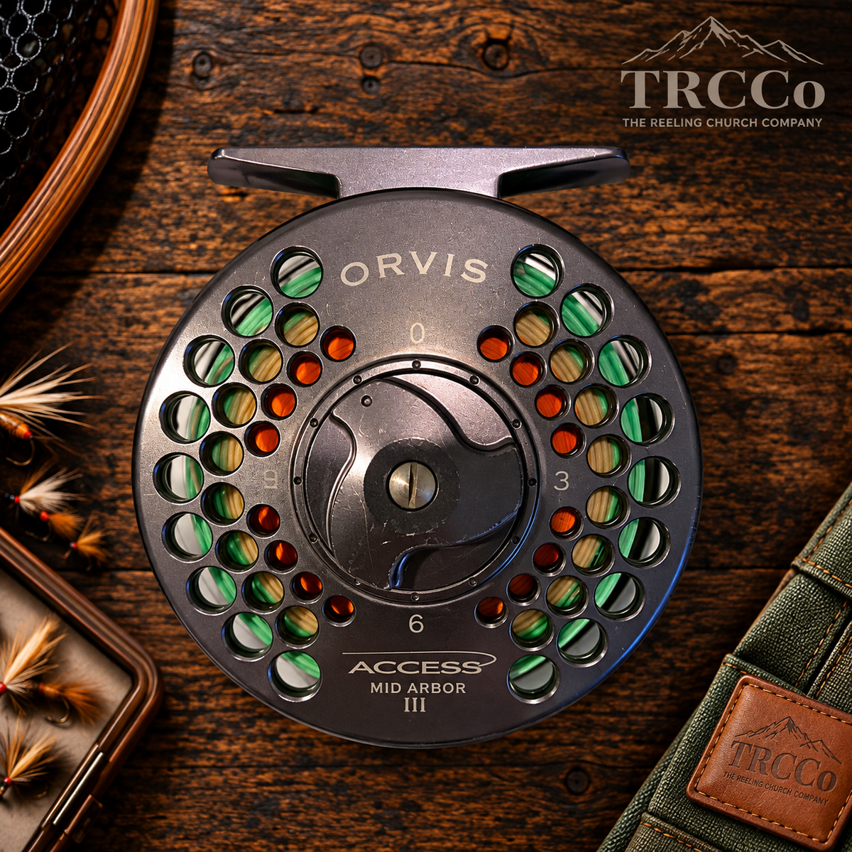 A close-up of a TRCCo Access Mid Arbor III fly reel by ORVIS, featuring a dark gray metal body with a multicolor patterned spool, displayed on a rustic wooden background with fishing accessories.