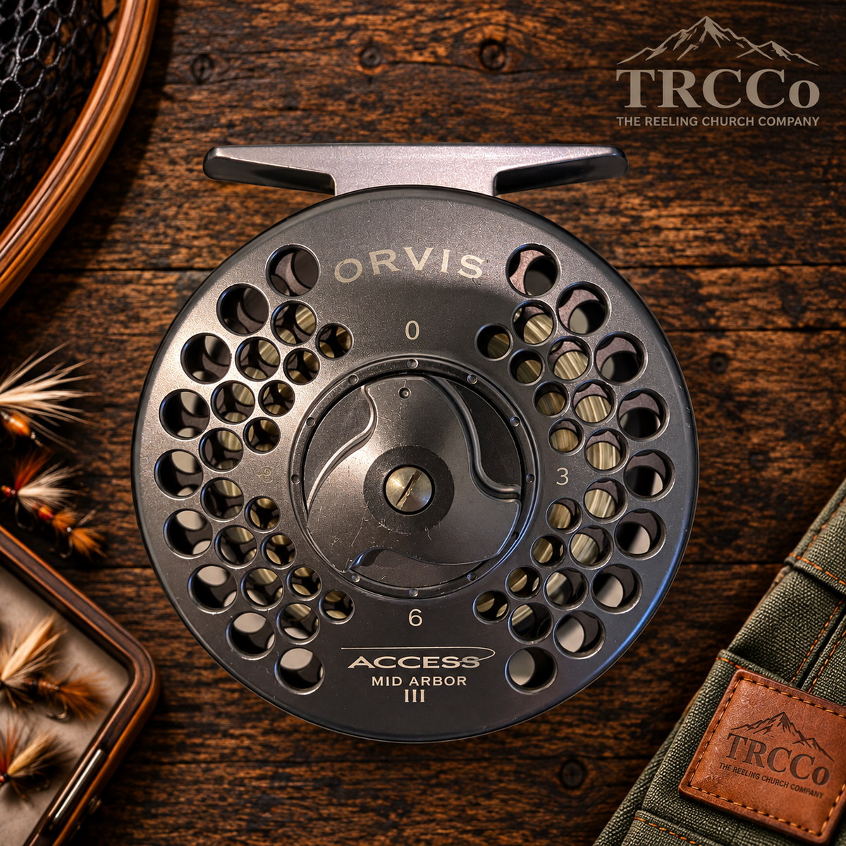 A close-up of a TRCco Access Mid-Arbor III fly fishing reel on a rustic dark wood background, surrounded by fishing accessories including a box of flies and a green strap with a brown leather patch.