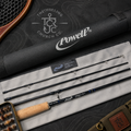Powell fly fishing rod set displayed on a light gray fabric surface with its black carrying case, shown in a styled flat-lay arrangement with a paddle and leather strap nearby. The rod is black with blue accents and a cork handle.