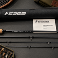 Sage fly fishing rod set laid out on a dark textured surface, including the rod sections, a black Sage-branded tube, a warranty card, and a small fly box. The rod has a cork handle and black shaft with silver hardware.