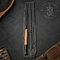 A fly fishing rod with a cork handle is displayed vertically in a black fabric rod roll, surrounded by flat-lay props including a wooden paddle, a fly box, and an olive green pouch with a leather patch. The background is a dark textured surface with the logo for 'The Reeling Church Co.